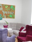 Pomieszczenie Dziennego Domu Pomocy. Dwa fotele w kolorach fioletowym i bordowym, narożna kanapa koloru szarego. Na białej ścianie tablica korkowa z napisem "Nasze miejsce spotkań"